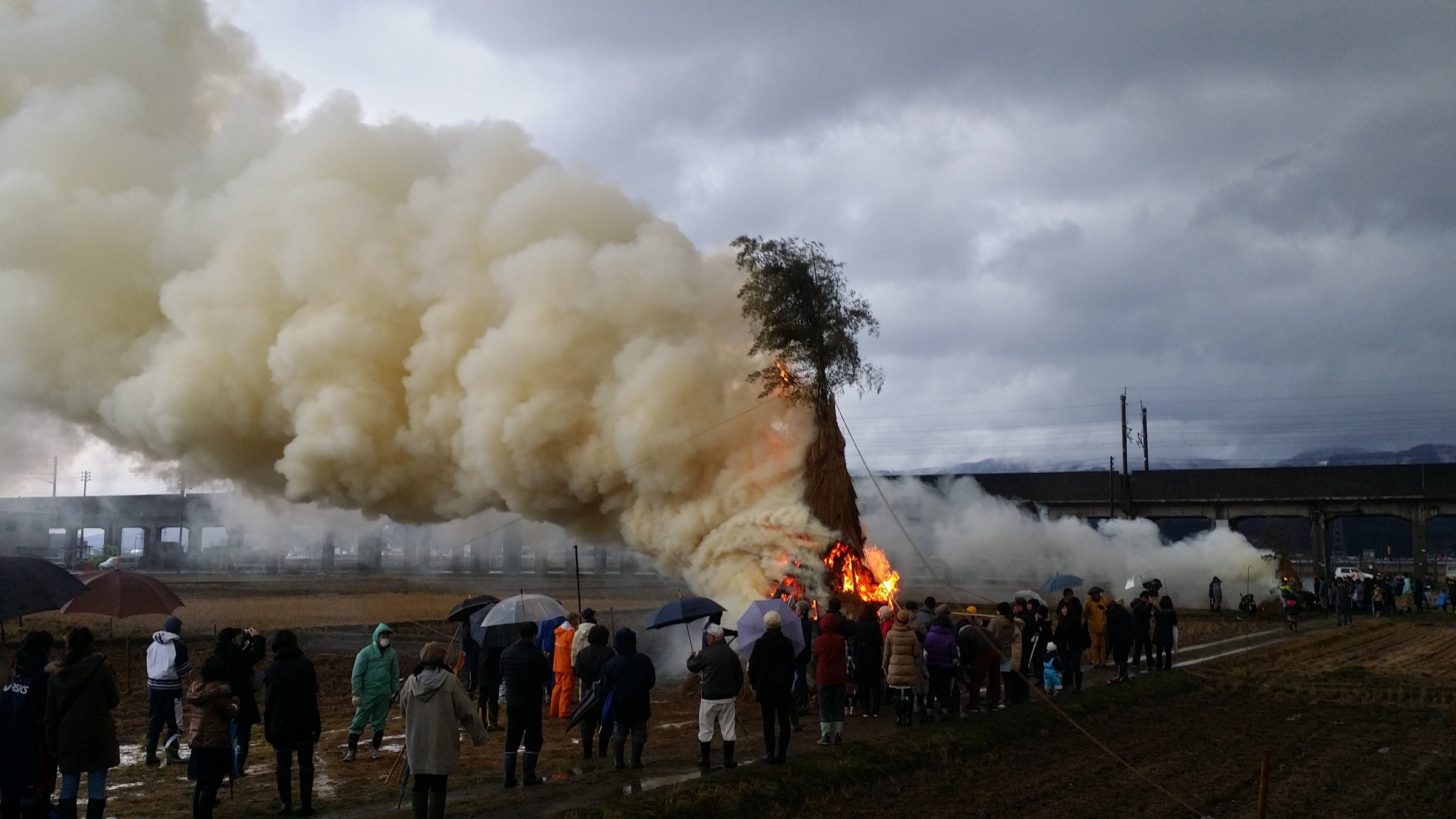 Alto cono di paglia con una cima verde da cui sale un'enorme nube di fumo, davanti spettatori con gli ombrelli